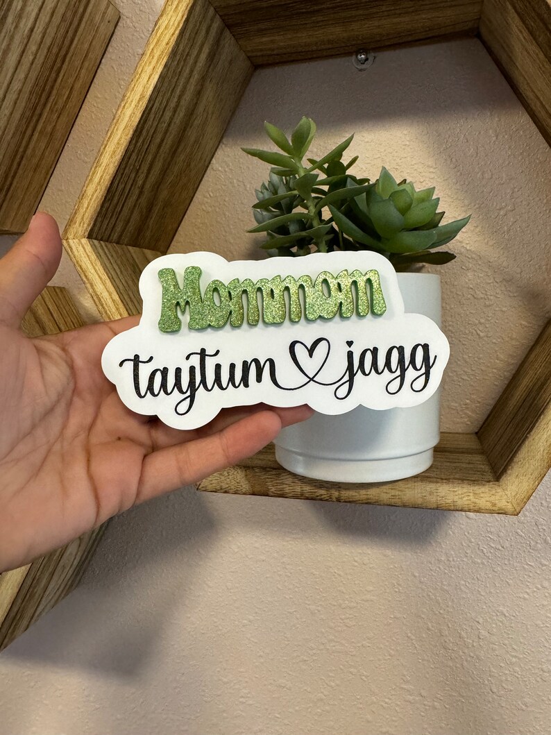 May include: A white sign with the words "Mommom Taytum & Jagg" in black lettering. The word "Mommom" is in green glitter.