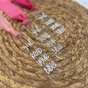 Peut inclure: Marque-pages transparents en acrylique avec le texte "girl READ YOUR BIBLE" en écriture blanche. Chaque marque-page est attaché à un ruban rose de teinte différente. Les marque-pages sont disposés sur une surface tissée brune.