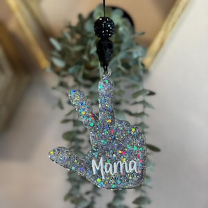 May include: A glittery peace sign ornament with the word "Mama" in white script. The ornament is silver with colorful flecks and is suspended from a black string with a black skull and beaded accent.