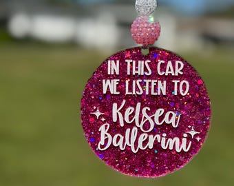 May include: A round, glittery pink car charm with the text "IN THIS CAR WE LISTEN TO Kelsea Ballerini" in white script. The charm is suspended by a white string and adorned with decorative beads.