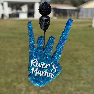May include: A blue glitter resin hand charm shaped in a rock-and-roll gesture with the white text River's Mama printed on the front. The charm hangs from a black cord featuring a black skull bead and a black textured spherical bead above it.