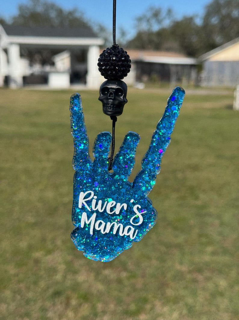 May include: A blue glitter resin hand charm shaped in a rock-and-roll gesture with the white text River's Mama printed on the front. The charm hangs from a black cord featuring a black skull bead and a black textured spherical bead above it.