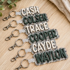 May include: Six rectangular keychains featuring a gray and black speckled pattern base. Each keychain has raised, three-dimensional lettering in either cream or dark green, displaying the names CASH, COLSEN, TRACE, COOPER, CAYDE, and WAYLON.