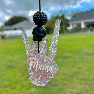 May include: A hanging ornament shaped like a skeleton hand making a rock-and-roll gesture, filled with iridescent glitter and the word Mama in white script. It features a black skull bead and a textured black bead on a black cord string.