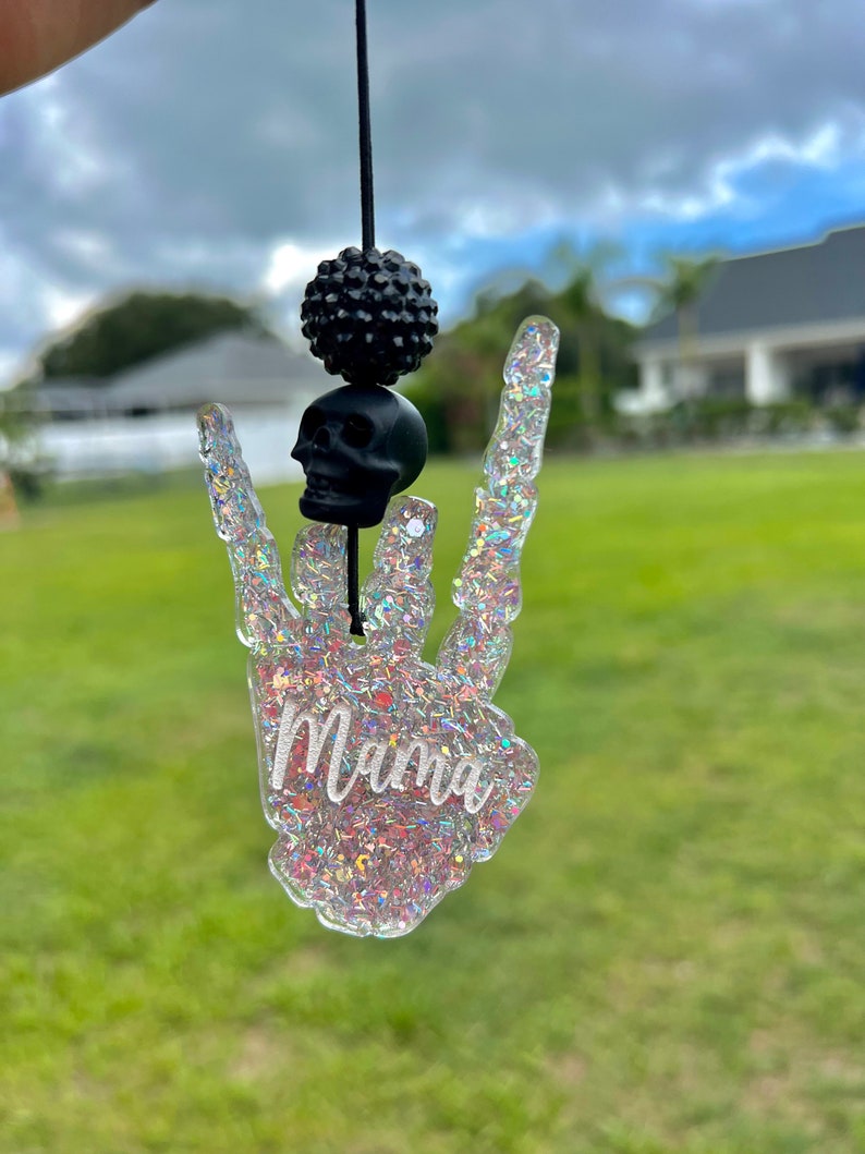 May include: A hanging ornament shaped like a skeleton hand making a rock-and-roll gesture, filled with iridescent glitter and the word Mama in white script. It features a black skull bead and a textured black bead on a black cord string.