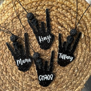 May include: Four black glittery skeleton hand ornaments shaped in a rock-on gesture, each featuring a black skull bead, a black textured bead, and a black cord. The ornaments are personalised with white script text reading Mama, Kenzi, Chaos, and Tiffany.