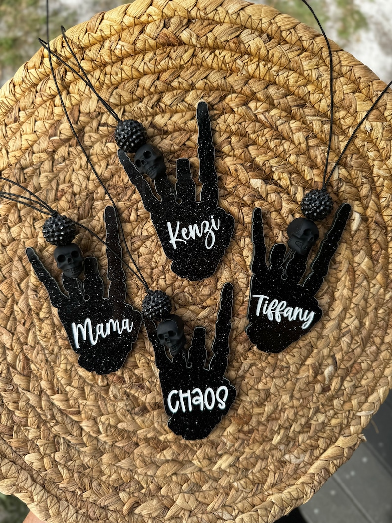 May include: Four black glittery skeleton hand ornaments shaped in a rock-on gesture, each featuring a black skull bead, a black textured bead, and a black cord. The ornaments are personalised with white script text reading Mama, Kenzi, Chaos, and Tiffany.
