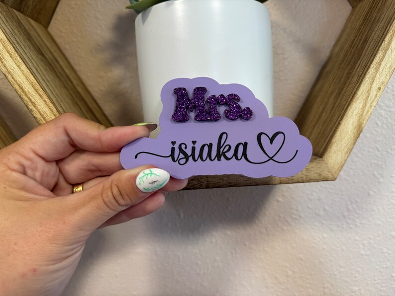 May include: Purple cloud-shaped wooden sign with the text "Mrs. isiaka" in black cursive lettering and a heart symbol. The word "Mrs." is in purple glitter.