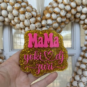 May include: Gold glitter sign with the words "Mama" in pink, a pink heart, and the words "yoki dj zuri" in pink cursive.