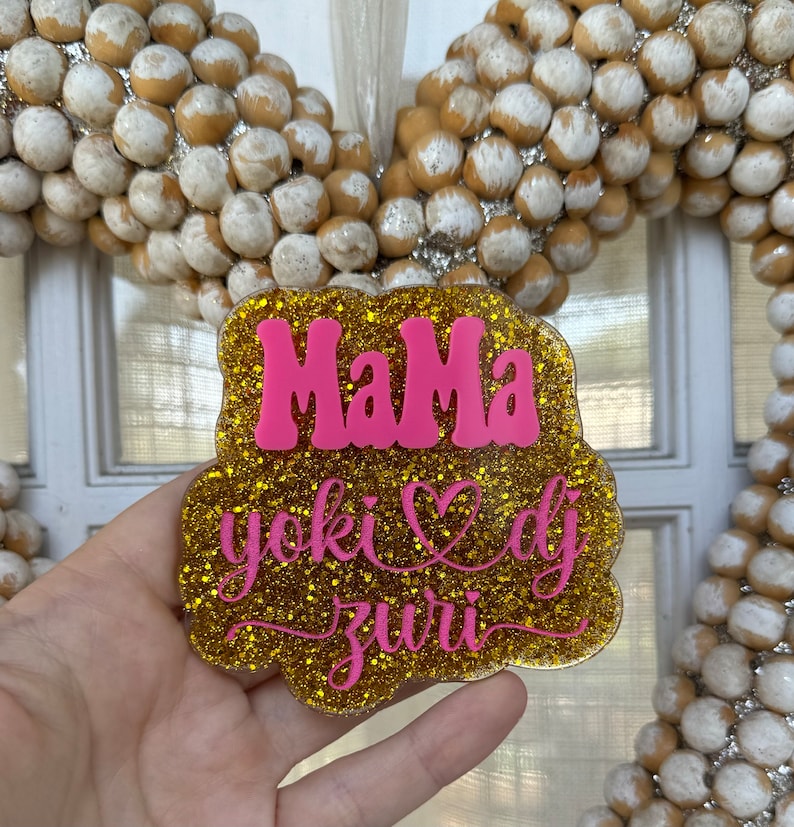 May include: Gold glitter sign with the words "Mama" in pink, a pink heart, and the words "yoki dj zuri" in pink cursive.