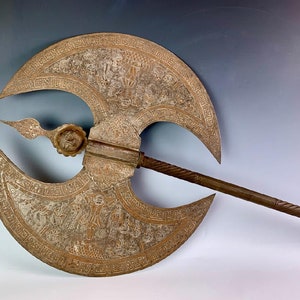 Puede incluir: Un hacha de batalla antigua y ornamentada con una hoja en forma de media luna. La hoja presenta intrincadas tallas e inscripciones. El mango es largo y retorcido, con un elemento decorativo cerca de la hoja. El hacha tiene una apariencia metálica desgastada.