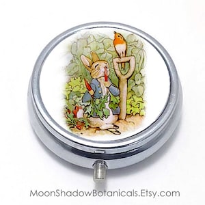 May include: A silver round pill box with a white lid featuring a vintage illustration of Peter Rabbit eating a carrot with a robin perched on a garden tool.