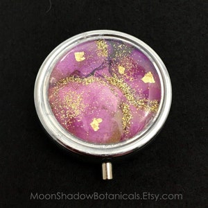May include: A round silver pill box with a hinged lid. The lid has a clear top with a pink and gold resin design. The resin design features gold glitter and gold leaf accents.