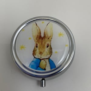 May include: A silver round pill box with a white lid featuring a cartoon image of Peter Rabbit in a blue jacket. The background is white with yellow stars.