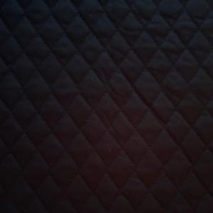 May include: A close-up of a black quilted fabric. The fabric has a diamond pattern created by stitched lines. The texture appears soft and the color is a deep, solid black. The lighting creates shadows that emphasize the pattern.