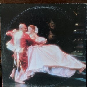 May include: A black and white photograph of a couple dancing. The woman is wearing a white dress and the man is wearing a red jacket. The couple are dancing in a ballroom setting.