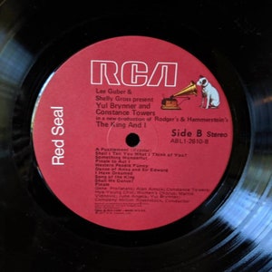 May include: Red Seal RCA vinyl record with a red label featuring a black and white illustration of a gramophone and a dog. The label text reads "Lee Guber & Shelly Gross present Yul Brynner and Constance Towers in a new production of Rodger's & Hammerstein's The King And I Side B Stereo ABL1-2610-B".