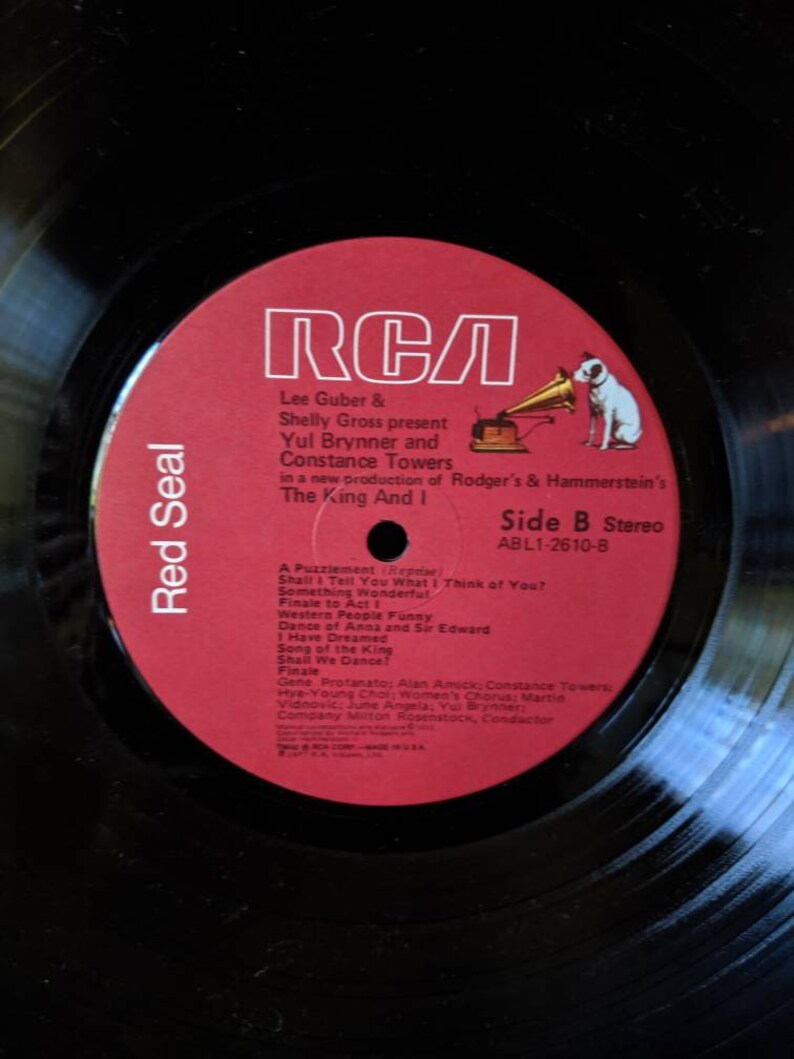 May include: Red Seal RCA vinyl record with a red label featuring a black and white illustration of a gramophone and a dog. The label text reads "Lee Guber & Shelly Gross present Yul Brynner and Constance Towers in a new production of Rodger's & Hammerstein's The King And I Side B Stereo ABL1-2610-B".