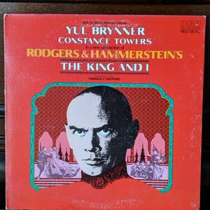 May include: A red vinyl record album cover featuring a black and white portrait of Yul Brynner as the King of Siam from the musical "The King and I". The album cover also features the title "The King and I" and the names of the actors Yul Brynner and Constance Towers.