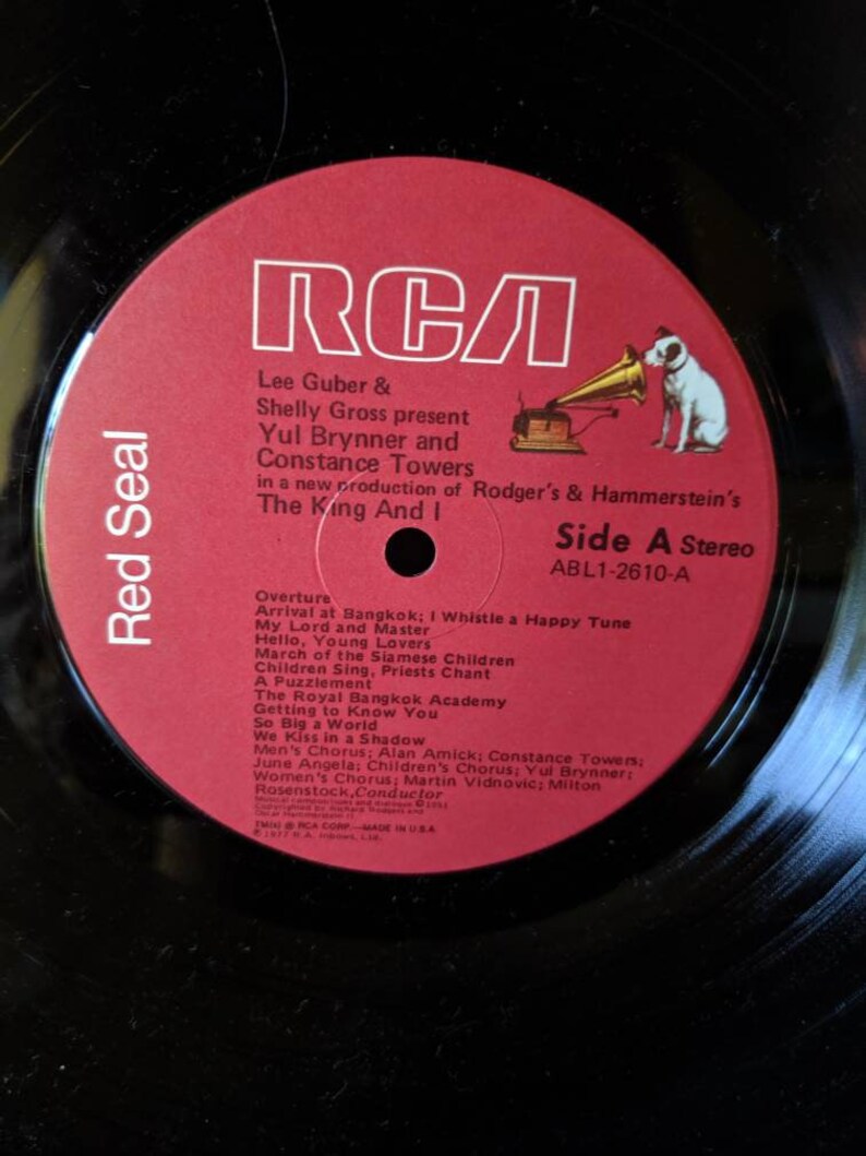 May include: A red and white vinyl record label featuring the RCA logo and the title "The King And I" in black text. The label also includes the names Yul Brynner and Constance Towers, and the song titles "Overture", "Arrival at Bangkok", "My Lord and Master", "Hello, Young Lovers", "March of the Siamese Children", "Children Sing, Priests Chant", "A Puzzlement", "The Royal Bangkok Academy", "Getting to Know You", "So Big a World", "We Kiss in a Shadow", "Men's Chorus", "June Angela", "Children's Chorus", "Women's Chorus", and "Milton Rosenstock, Conductor".