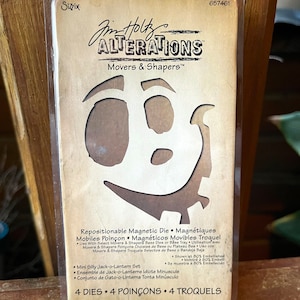 Könnte beinhalten: Nahaufnahme einer Verpackung für einen Satz von vier repositionierbaren magnetischen Stanzformen zum Basteln. Die Verpackung ist braun und zeigt ein Comic-Gesicht mit einem Zahnrad. Der Text auf der Verpackung lautet "Jim Holtz Alterations Movers & Shapers" und "4 Dies • 4 Poinçons • 4 Troquels".