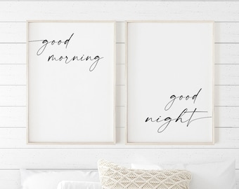 May include: Two framed prints with white backgrounds and black calligraphy. The left print says "good morning" and the right print says "good night". The prints are hanging on a white shiplap wall above a bed with a white and gray striped duvet cover and a white throw blanket.