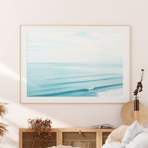 May include: A white bed with a light brown throw blanket and white pillows. The bed is in a bedroom with a wooden cabinet and a framed photograph of a beach scene above the cabinet.