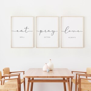 May include: Three framed prints with the text "eat well, pray often, love always" hanging on a white wall above a wooden table with two chairs.