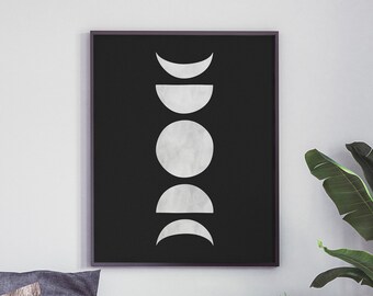 May include: A black and white minimalist moon phase print in a black frame hanging on a white wall above a gray bed with a black throw blanket. A white nightstand with a drawer is to the right of the bed.