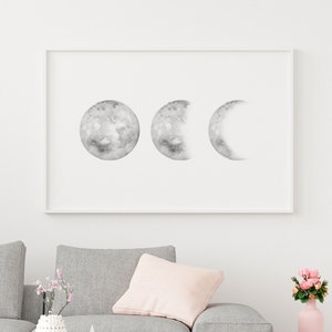 May include: A framed black and white print of three moons in different phases, hanging on a white wall above a gray sofa with a pink throw pillow. A vase with pink flowers and a white ceramic vase with a geometric design are on a wooden coffee table in front of the sofa.