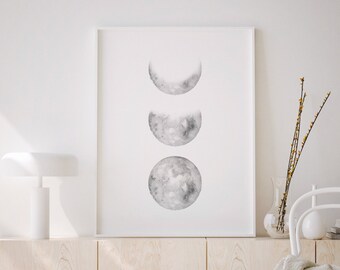 May include: A white framed print with a watercolor illustration of three moon phases in shades of gray. The print is hanging on a white wall above a light wood cabinet with a white lamp and a white chair with a white throw blanket.