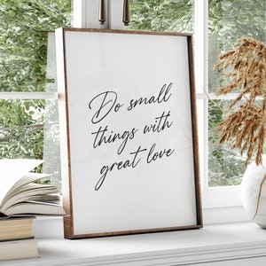 Puede incluir: Una impresión enmarcada con fondo blanco y texto negro que dice "Do small things with great love". La impresión está sobre una repisa blanca con un libro y un jarrón con flores secas.