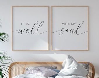 May include: Two framed prints with the text "It is well" and "With my soul" hanging on a white wall above a bed with white linens and a wooden headboard. The bed is in a bedroom with a wooden floor and a plant in a woven basket on the floor.