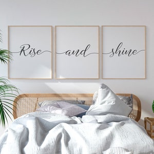 Op de afbeelding: Een wit bed met een lichtgrijs linnen dekbed en een houten hoofdeinde met geweven rieten details. Het bed is gestyled met kussens en een deken. Drie ingelijste prints met de tekst "Rise and shine" hangen aan de muur boven het bed.