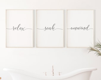 May include: Three framed prints with the words "relax", "soak", and "unwind" in a white bathroom with a white bathtub and a wooden stool.