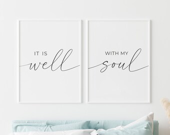 May include: Two white framed prints with black calligraphy. The left print says "It is well" and the right print says "With my soul". The prints are hanging on a white wall above a bed with light blue bedding.