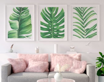 Könnte beinhalten: Ein graues Sofa mit drei rosa Kissen und einem Glas-Couchtisch davor. An der Wand hinter dem Sofa hängen drei gerahmte Aquarellbilder von tropischen Blättern.