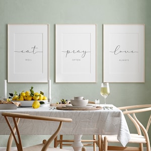 May include: Three framed prints with white backgrounds and black text on a light green wall. The prints read "eat well", "pray often", and "love always". The prints are hanging above a dining table with a white tablecloth and two wooden chairs.