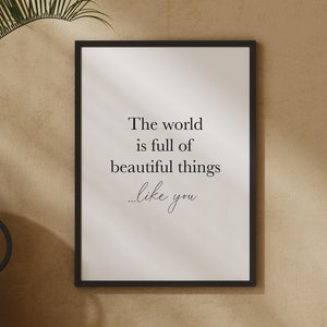 May include: A black framed print with the text "The world is full of beautiful things...like you" hanging on a beige wall. A green plant, a black vase, a woven vase, and a wooden table are in the foreground.