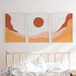 May include: Three abstract landscape prints in a minimalist style with a white background, orange and red hills, and a red circle in the center print. The prints are hanging above a wooden bed frame with white bedding.