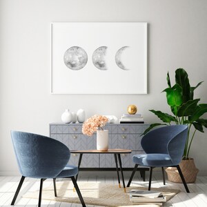 May include: Two blue chairs with black legs and a wooden table with a gray cabinet in the background. A framed print of a moon phases design hangs on the wall above the cabinet. A potted plant is on the right side of the image.