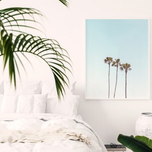 May include: A framed print of three palm trees against a pale blue sky. The print is hanging on a white wall in a bedroom setting.