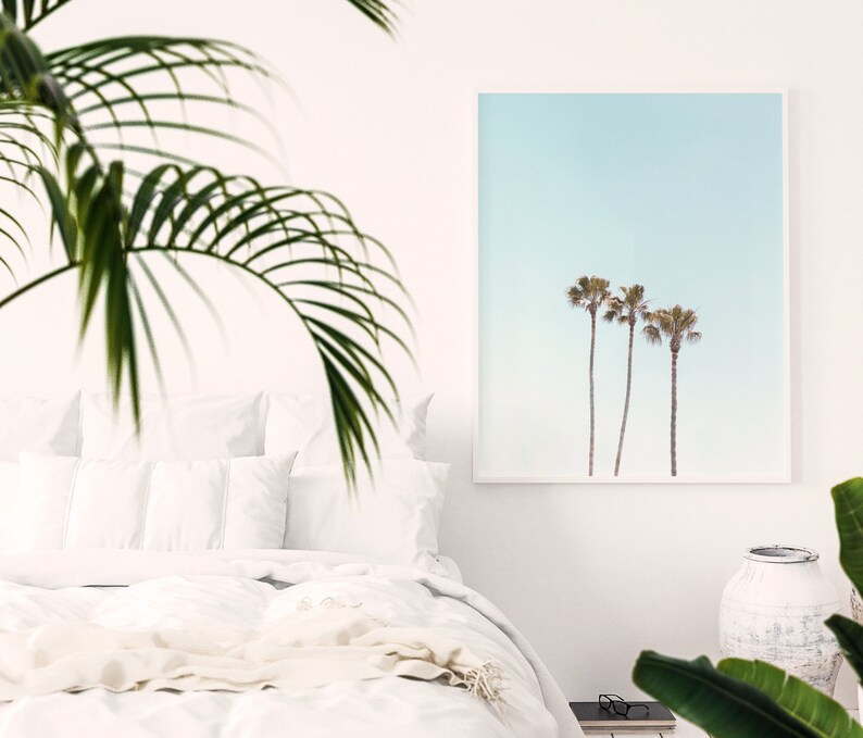 May include: A framed print of three palm trees against a pale blue sky. The print is hanging on a white wall in a bedroom setting.