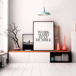 May include: A white shelf unit with a black top and a black shelf on the bottom. The shelf unit has two white drawers and a black shelf with books on it. There are two black framed prints on the wall above the shelf unit. The print on the left is a black and white photo of a person in the water. The print on the right has the text "WORK HARD STAY HUMBLE". There is a light blue pendant light hanging from the ceiling above the prints.
