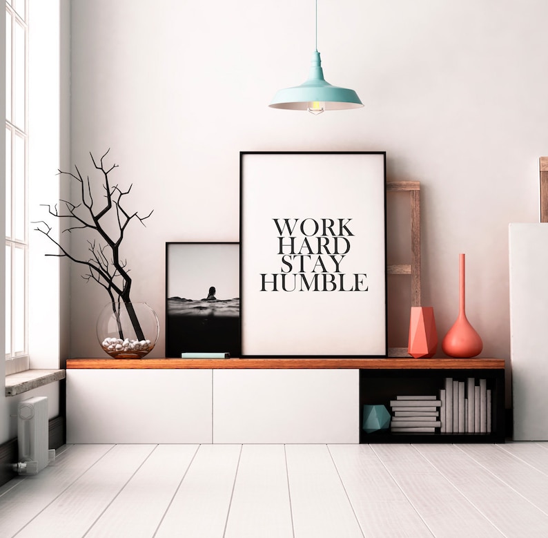 May include: A white shelf unit with a black top and a black shelf on the bottom. The shelf unit has two white drawers and a black shelf with books on it. There are two black framed prints on the wall above the shelf unit. The print on the left is a black and white photo of a person in the water. The print on the right has the text "WORK HARD STAY HUMBLE". There is a light blue pendant light hanging from the ceiling above the prints.