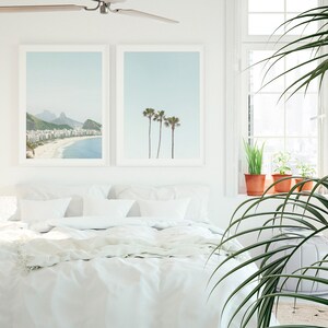 May include: A white bed with white bedding and two framed prints on the wall behind it. The print on the left shows a beach scene with a mountain in the background. The print on the right shows three palm trees against a blue sky. A large green plant is in the foreground.
