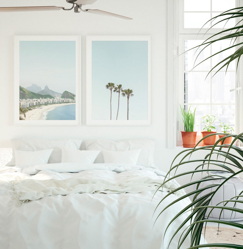 May include: A white bed with white bedding and two framed prints on the wall behind it. The print on the left shows a beach scene with a mountain in the background. The print on the right shows three palm trees against a blue sky. A large green plant is in the foreground.