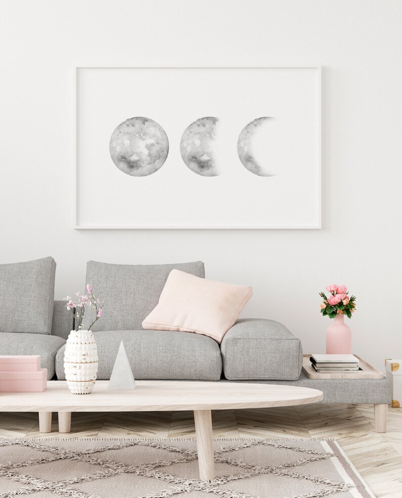 May include: A framed black and white print of three moons in different phases, hanging on a white wall above a gray sofa with a pink throw pillow. A vase with pink flowers and a white ceramic vase with a geometric design are on a wooden coffee table in front of the sofa.