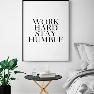 May include: Black and white framed print with the text "WORK HARD STAY HUMBLE" hanging on a white wall above a black side table with a glass of water and books. The table has a wooden base and is in front of a gray bed with white bedding.