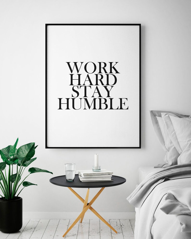 May include: Black and white framed print with the text "WORK HARD STAY HUMBLE" hanging on a white wall above a black side table with a glass of water and books. The table has a wooden base and is in front of a gray bed with white bedding.
